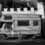 carado - A vintage camper van parked beside a brick wall, showcasing abandonment and decay in a mono